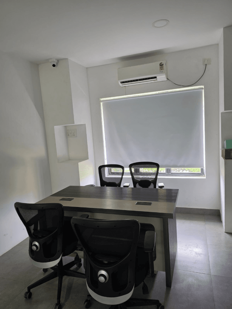 Small meeting room with a wooden table, four chairs, an air conditioner, and a window with a roller blind